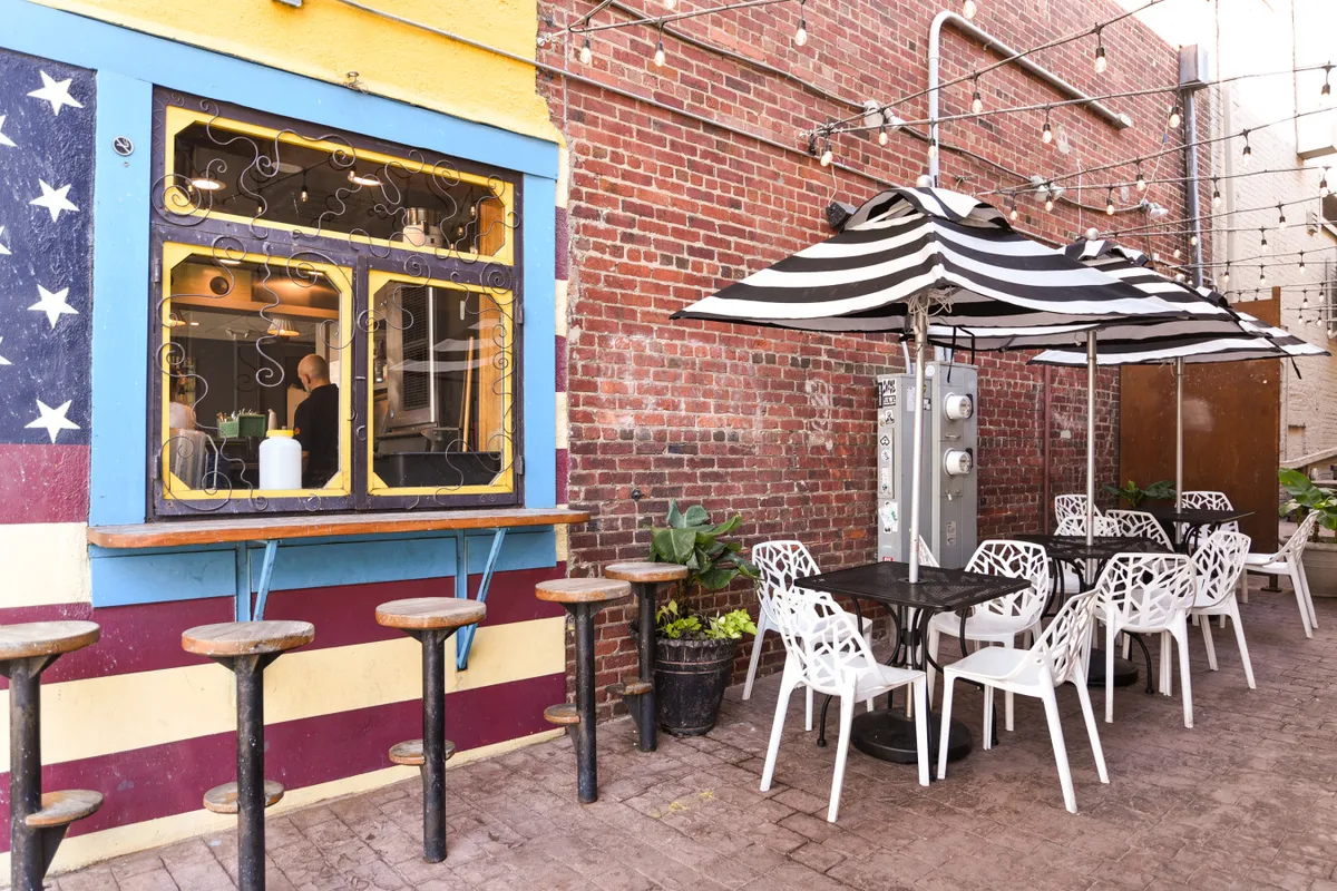 A colorful and inviting outdoor dining area, highlighted by a vibrant window with artistic patterns, striped umbrellas, and a casual seating space. The scene is bright and bustling, suggesting a place where locals and visitors gather to eat, socialize, and enjoy the creative ambiance.