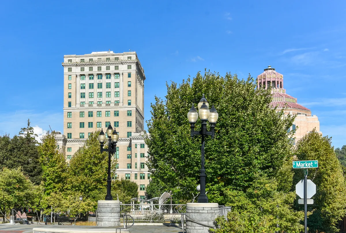 A lively Asheville streetscape featuring historic buildings, leafy trees, and classic street lamps. The scene is vibrant and inviting, suggesting an energetic urban environment ideal for walking and exploration.