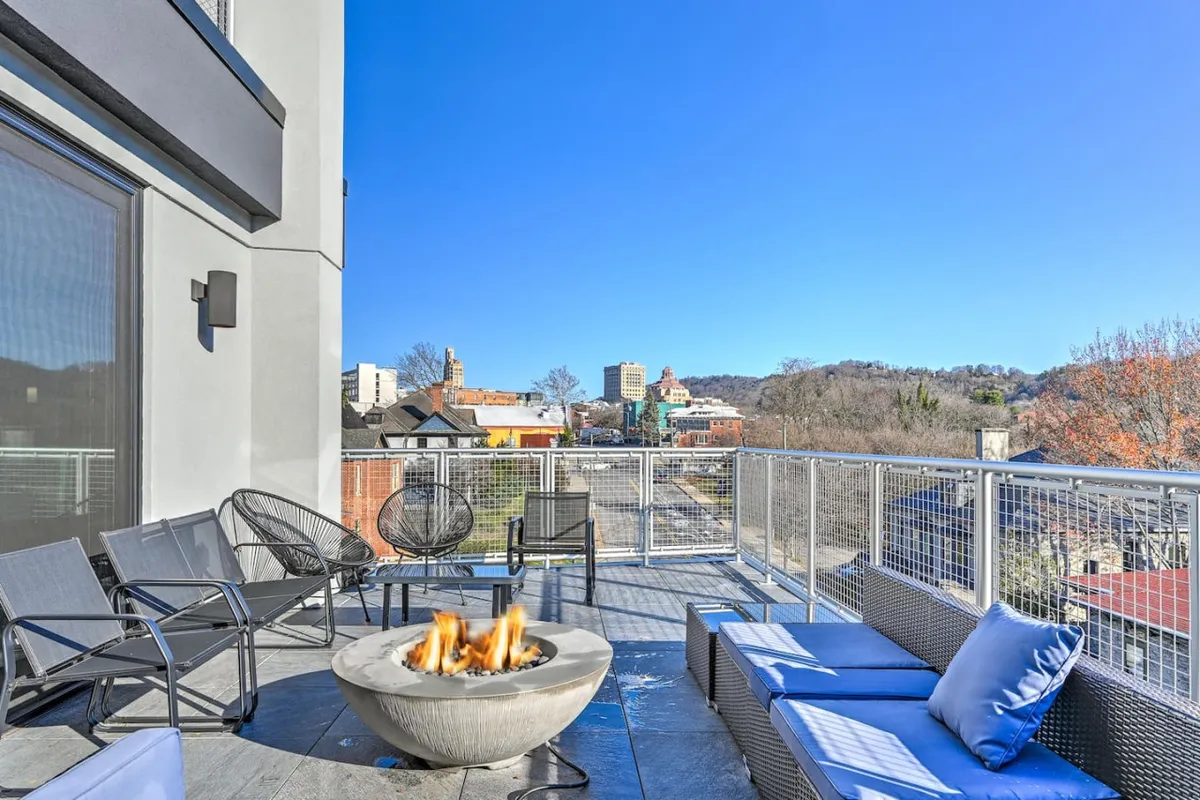 The image showcases an inviting outdoor seating area centered around a glowing fire pit, offering sweeping views of Asheville's twinkling city lights and the distant Blue Ridge Mountains. Comfortable chairs are arranged for socializing, and the spacious balcony setting is perfect for enjoying sunsets, barbecues, or stargazing beneath the open sky.