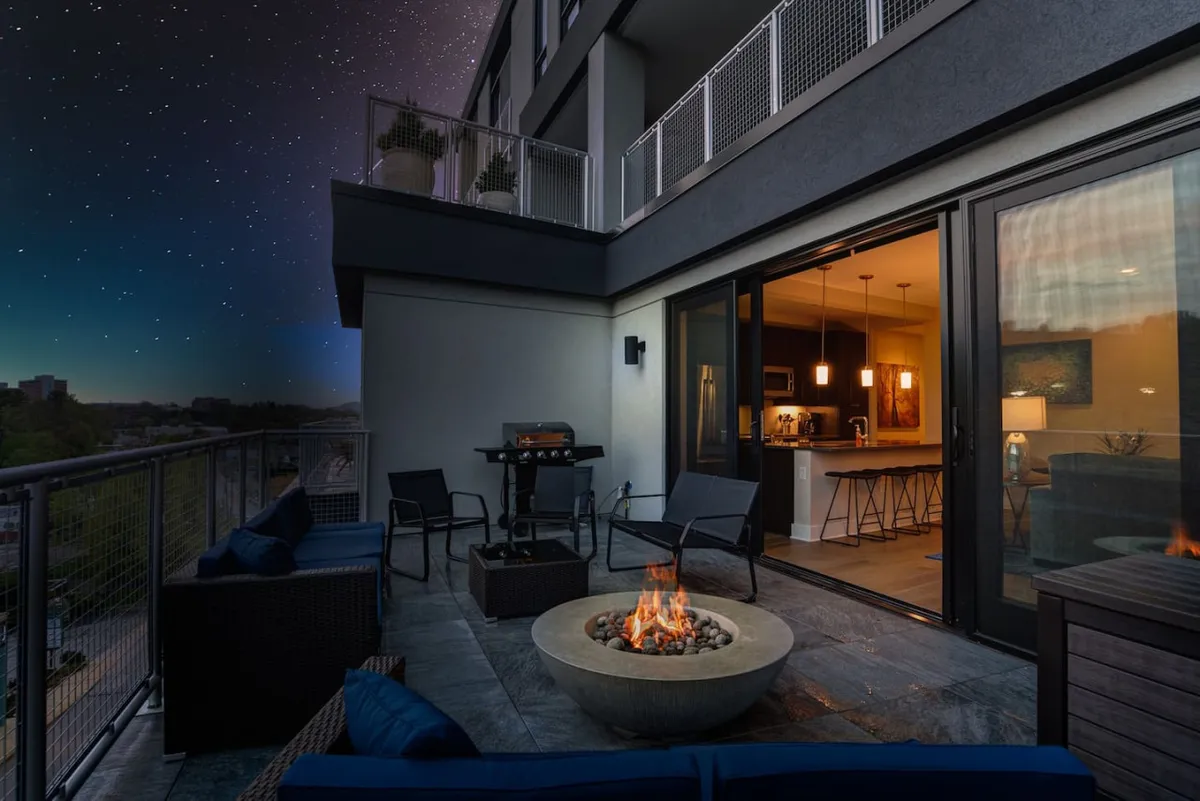 A cozy balcony scene with outdoor seating, a fire pit, grill, and an open door connecting to the kitchen. The space evokes intimacy and relaxation, with the warmth of the fire and the convenience of direct kitchen access.