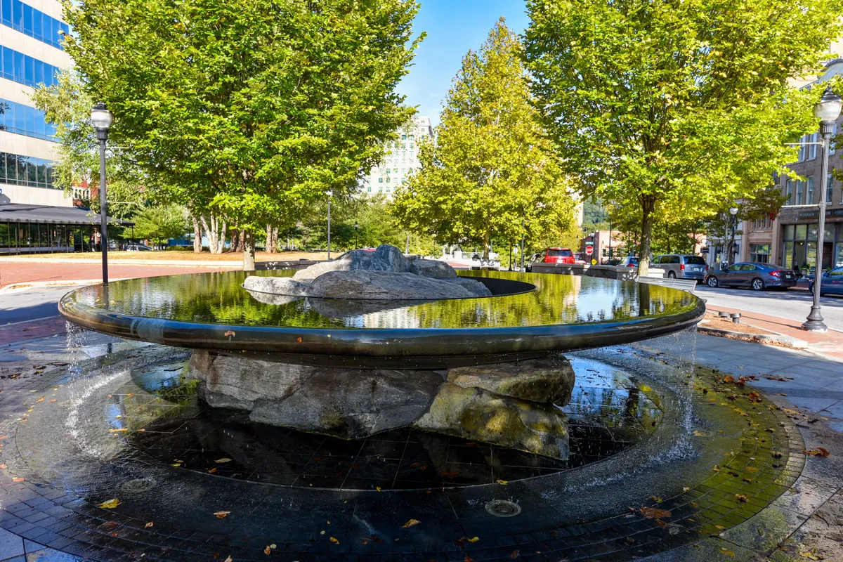 A tranquil city scene of a picturesque fountain surrounded by lush trees and reflected water. A sidewalk meanders nearby, inviting leisurely strolls and moments of reflection, with soft evening light enhancing the romantic atmosphere.
