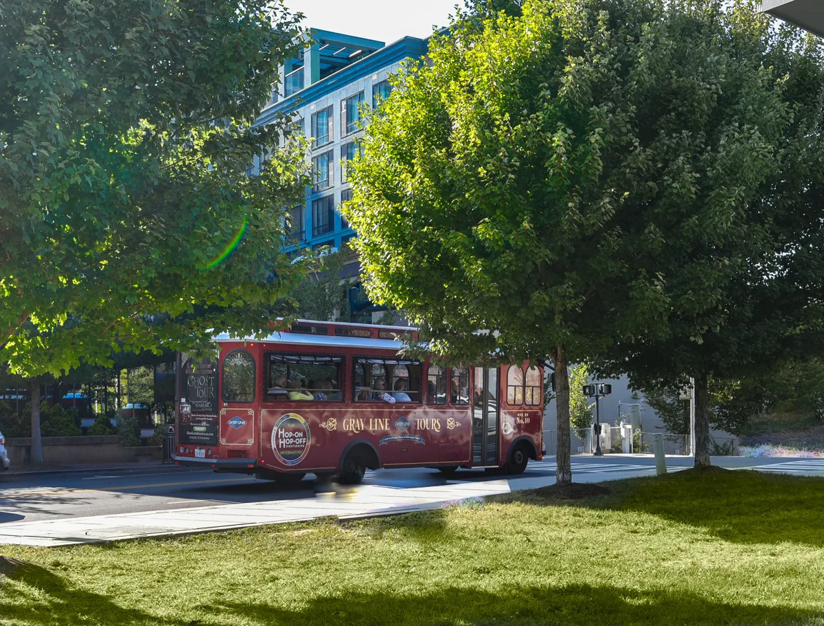 A red trolley bus winds past green trees and a modern downtown building, illustrating an active cityscape. The presence of the trolley suggests easy access to local attractions and a spirit of exploration, while the greenery adds to the city's charm.