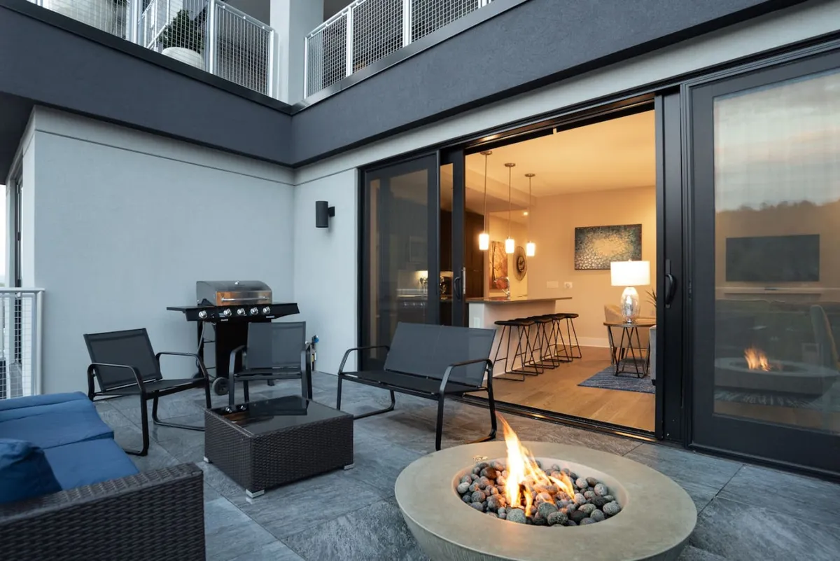 An inviting, expansive balcony featuring an outdoor fire pit, plush seating, grill, and sliding glass doors that lead seamlessly into the interior. The area is softly lit for evening gatherings, surrounded by city lights and distant mountain silhouettes.