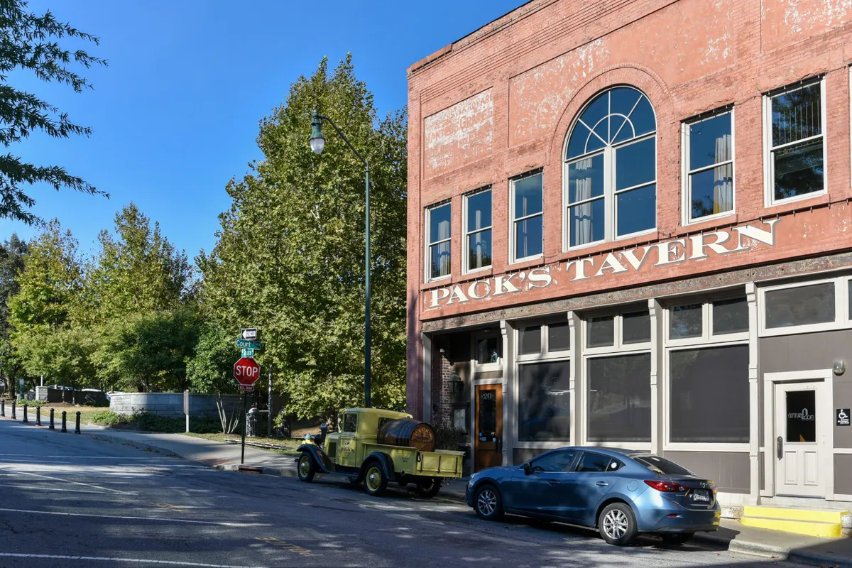 A lively downtown street scene featuring a historic brick building with large windows, a classic yellow truck parked in front, and clear street signage. The area appears active and bustling, suggesting nearby dining and social options, with people potentially gathering for live music or local events.