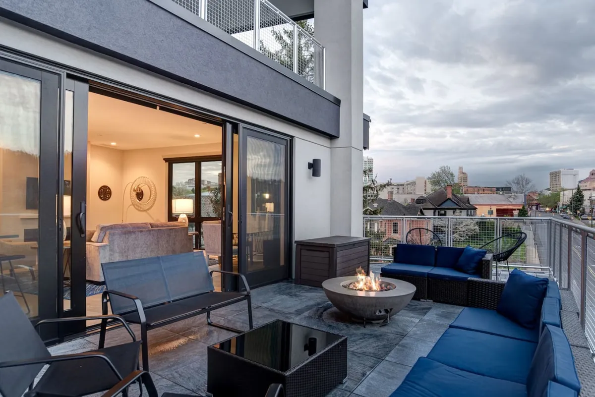 A stylish balcony overlooking the city, featuring a circular fire pit surrounded by modern outdoor furniture. The city skyline is visible beyond, enhancing the ambiance for social gatherings or quiet moments by the fire.