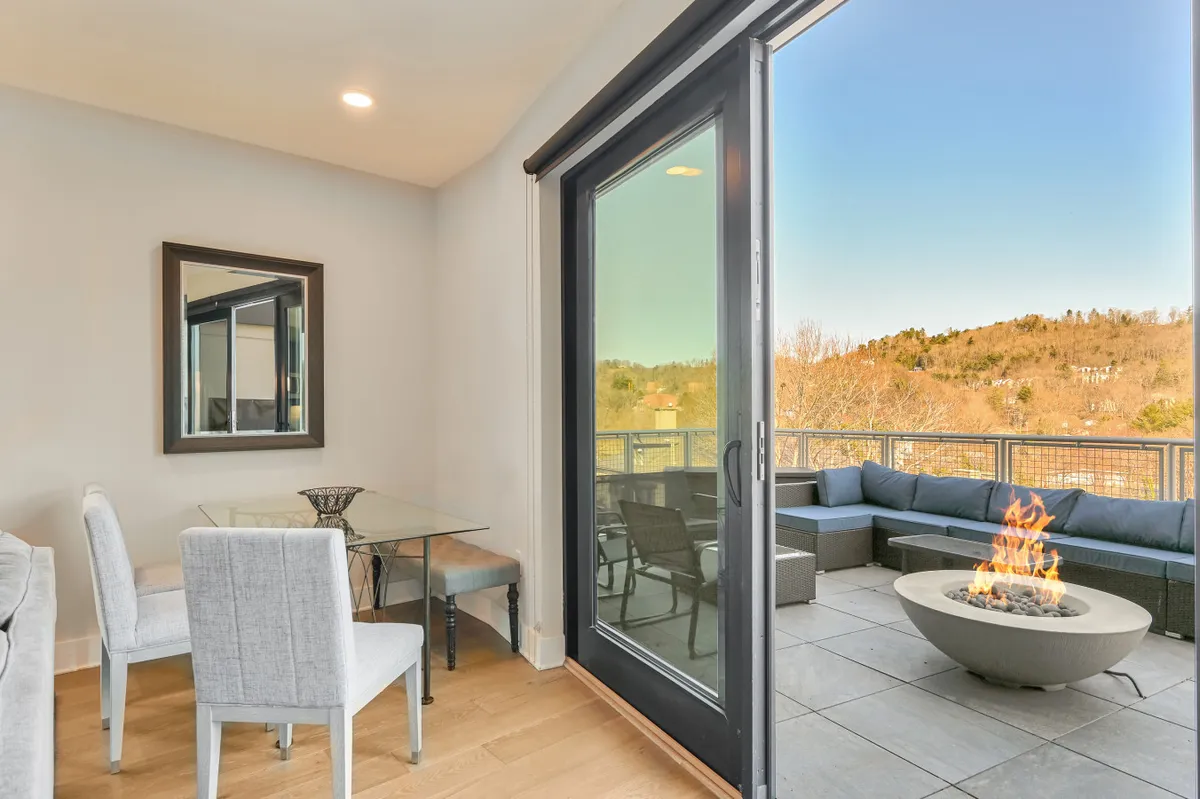 The image shows a welcoming interior dining area with a large dining table, a cozy fire pit, and large glass doors opening to a balcony. The view of the hills beyond the glass creates a seamless connection between indoor and outdoor spaces, enhancing the sense of memorable meals and gatherings.