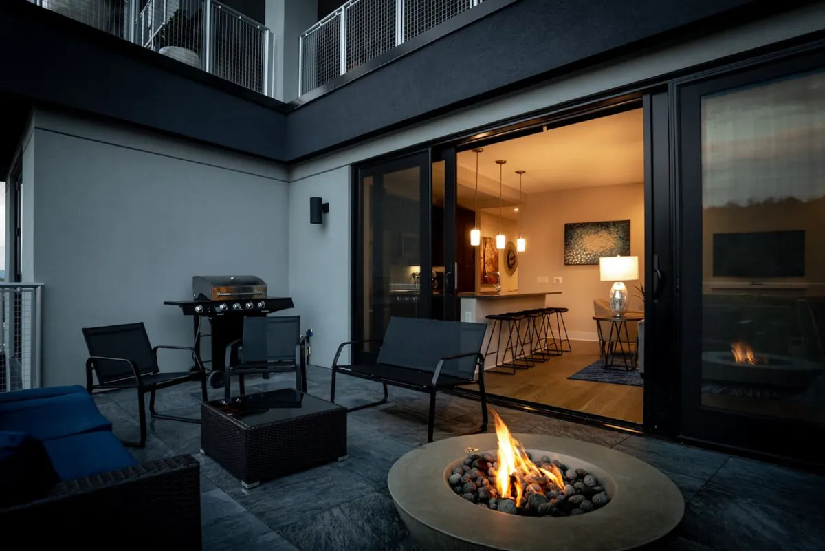 An inviting exterior scene featuring a fire pit with lively flames, plush outdoor lounge seating, a grill, and large sliding glass doors that seamlessly connect the patio to the interior. The warm evening light and comfortable setup make it ideal for both relaxing and entertaining.